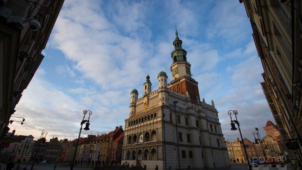 Ratusz w Starym Rynku widoczny od dołu, na zdjęciu uchwycono pogodne, pełne puszystych chmur niebo oraz otaczające ratusz zabytkowe kamienice i latarnie.