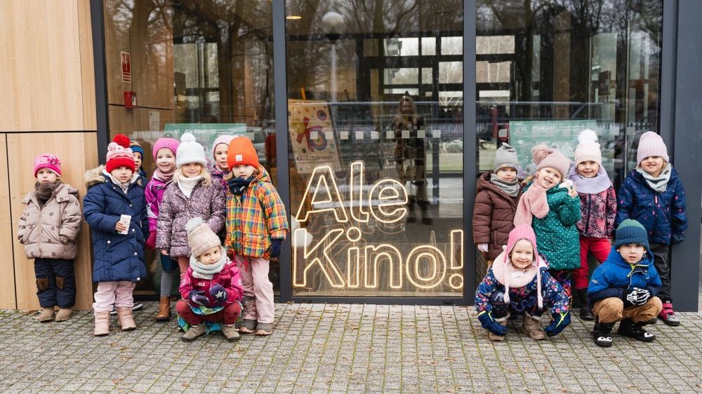 Przed wejściem do kina widać grupkę dzieci w kolorowych kurtkach i czapkach, uśmiechają się i machają do fotografki, której odbicie jest delikatnie widoczne w szklanych drzwiach.