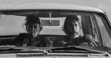 Czarno-białe zdjęcie mężczyzny prowadzącego samochód, obok niego pies. Na tylnym siedzeniu siedzi starsza kobieta.