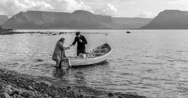 Czarno-biały kadr filmowy. Mężczyzna w czarnym stroju stoi na łódce, obok niego, w wodzie, stoi drugi mężczyzna. Za nimi fiord.