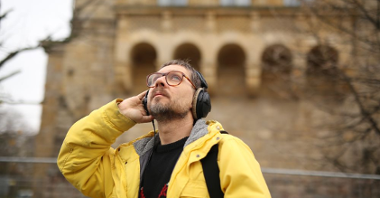 Mężczyzna w żółtej kurtce i okularach słucha nagrania w słuchawkach. Za nim budynek Centrum Kultury Zamek.