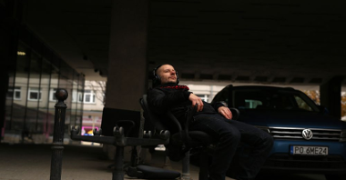 Mężczyzna w słuchawkach siedzi na krześle na ulicy. Za nim stoi niebieski samochód.