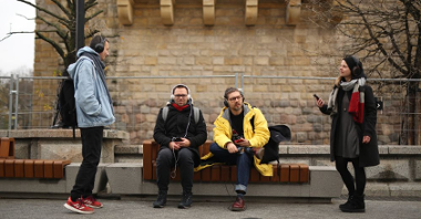 Czworo młodych ludzi stoi lub siedzi przed CK Zamek w Poznaniu. Słuchają nagrania audio na swoich telefonach.