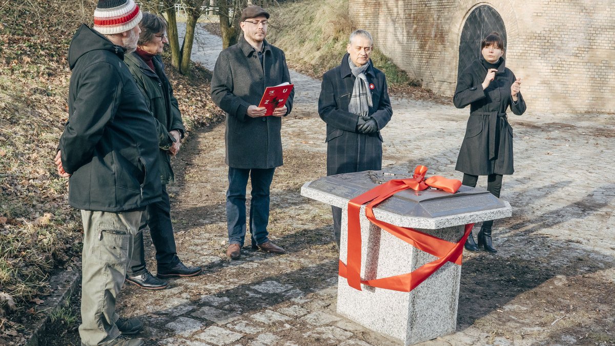 Czterej mężczyźni i jedna kobieta stoją wokół makiety twierdzy, kobieta tłumaczy wypowiedzi na język migowy