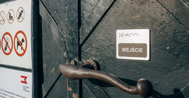 Metalowe drzwi i klamka, na nich niewielka tabliczka z napisem wejście i z wypukłymi kropkami