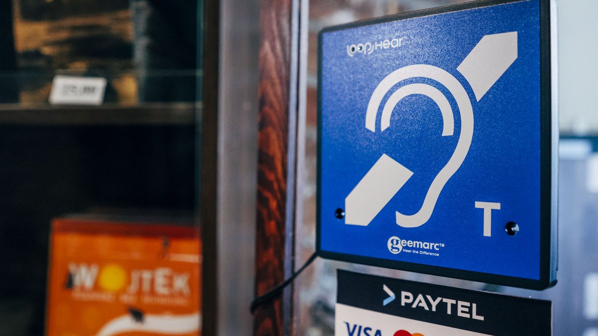 Oznaczenie pętli indukcyjnej na szklanej szybie kasy muzuealnej, obok oznaczenia kart płatniczych