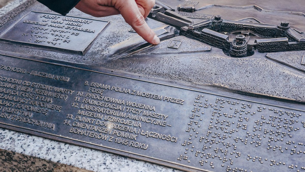 Ręka ludzka spoczywająca na metalowej konstrukcji makiety, na niej widoczne budynki dawnych koszar