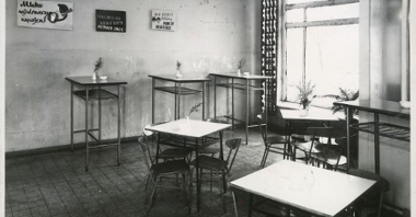 Czarno-białe zdjęcie pomieszczenia z kwadratowymi stolikami i krzesłami. Na ścianie blaszane tabliczki z komunikatami.