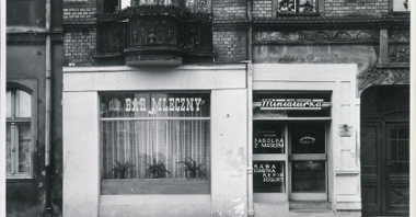 Czarno-białe zdjęcie witryny z napisami w różnych czcionkach. Nad witryną ceglana ściana i ozdobny, rzeźbiony balkon.