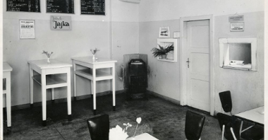 Sala z białymi stolikami i czarnymi krzesłami. W ścianie otwarte okienko do wydawania potraw.