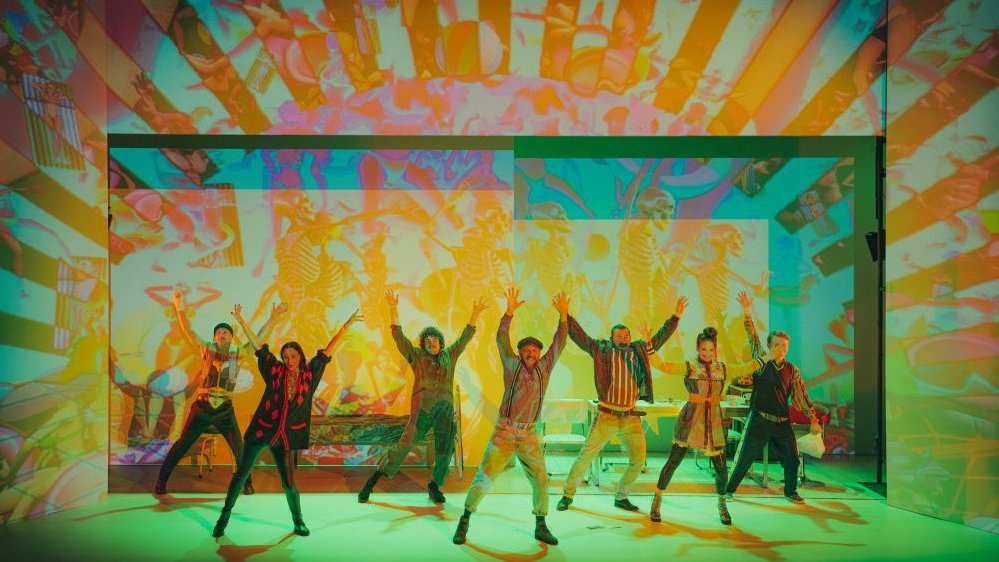 Grupa aktorów tańczy na scenie. Za nimi, na ścianie, pomarańczowo-żółte wizualizacje.