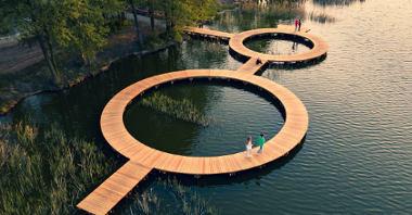 Wizualizacja drewnianego pomostu na wodzie. Pomost ma kształt dwóch połączonych okręgów, chodzą po nim ludzie.