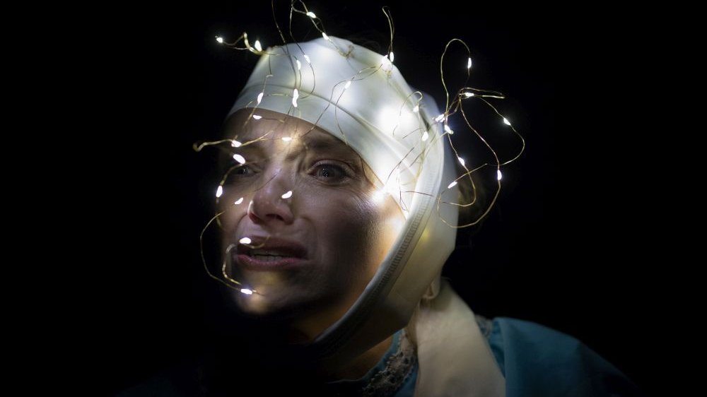 Kobieta w białym bandażu na głowie i z lampkami choinkowymi na nim robi zbolałą minę.