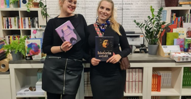 Dwie blondynki pozują ze swoimi książkami w rękach opierając się o ladę księgarni. Uśmiechają się szeroko.