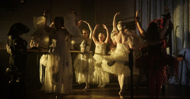 Tancerze baletowi, głównie w białych strojach, tańczą unosząc ręce w górę.