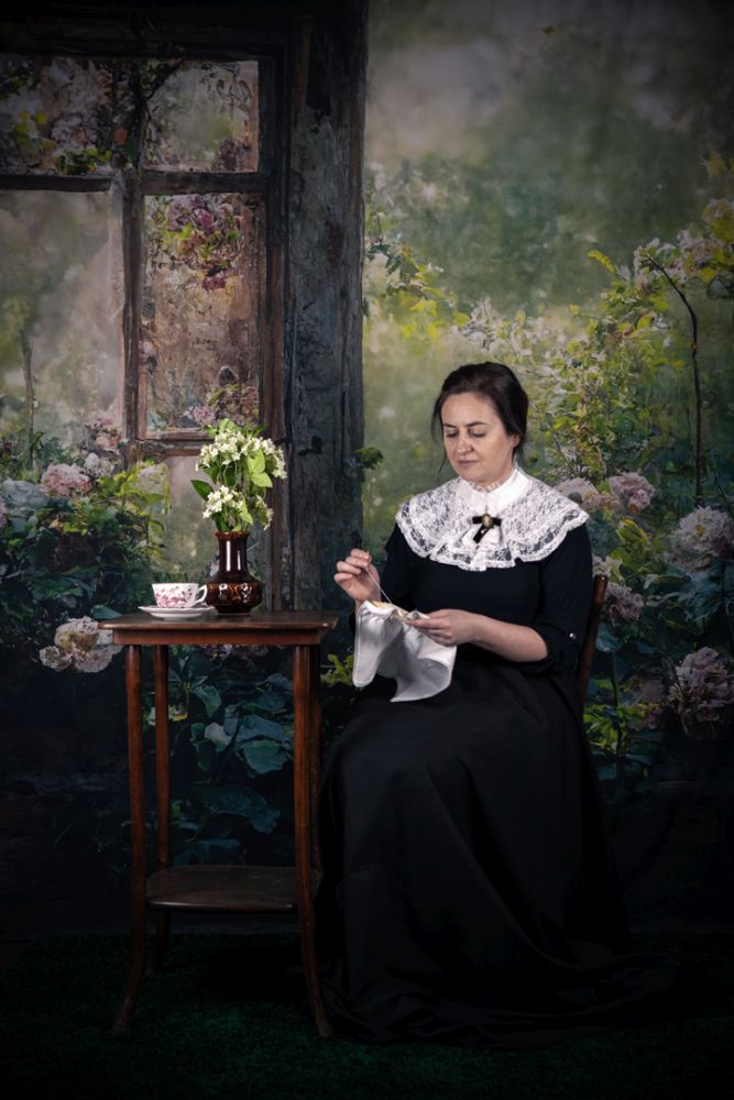 Kobieta w XIX-wiecznym stroju z dużym koronkowym kołnierzem wyszywa bialą chusteczkę siedząc przy stoliku. Na stoliku stoi wazon z bzem i porcelanowa filiżanka na spodku. - grafika artykułu