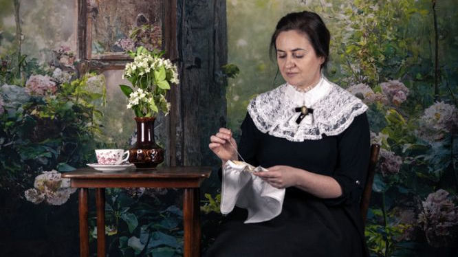 Kobieta w XIX-wiecznym stroju z dużym koronkowym kołnierzem wyszywa bialą chusteczkę siedząc przy stoliku. Na stoliku stoi wazon z bzem i porcelanowa filiżanka na spodku.