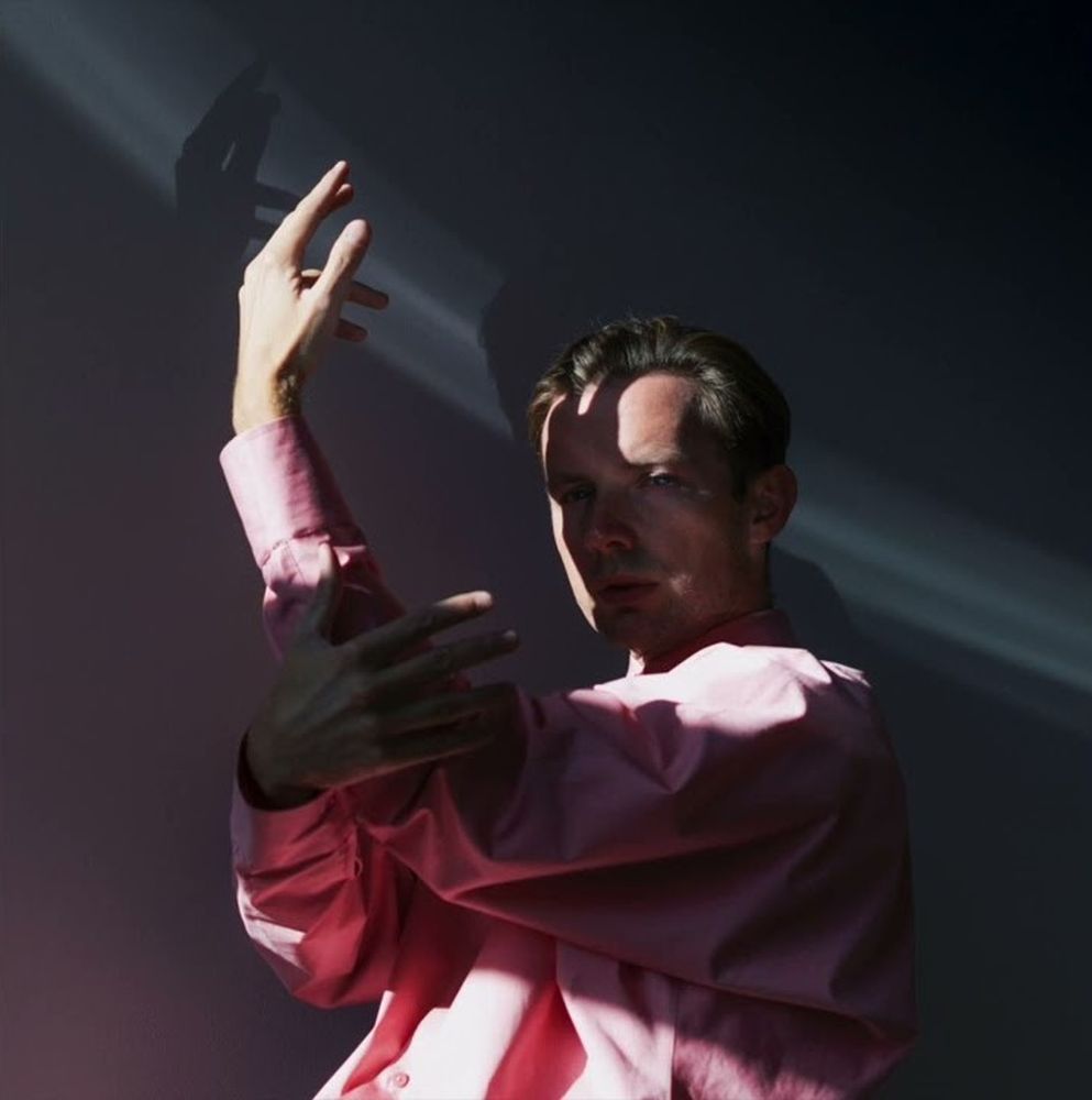 Mężczyzna w różowej koszuli stoi skryty w cieniu, artystycznie układa w powietrzu ręce. - grafika artykułu