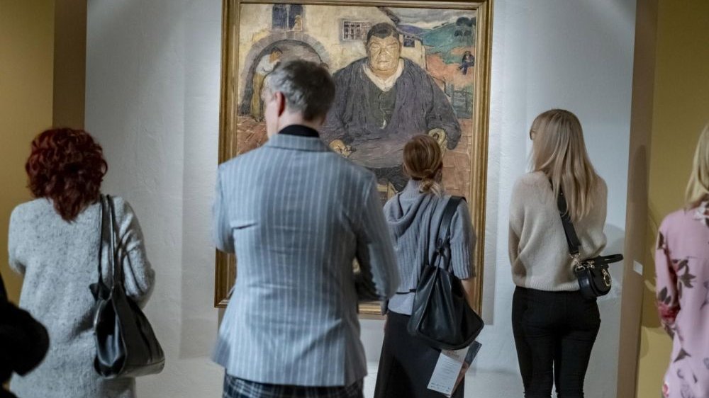 Ludzie na wystawie oglądają obraz przedstawiający mężczyznę, stoją tyłem do obiektywu.