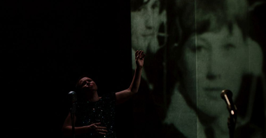 Kobieta w ciemności wygina ciało przy mirofonie. Na ekranie projekcja starych zdjęć.