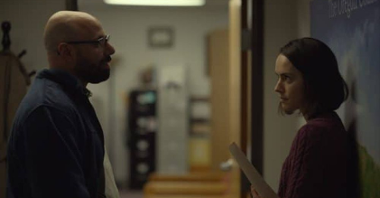 Łysy mężczyzna w okularach rozmawia z dziewczyną w krótkich włosach na korytarzu biura.
