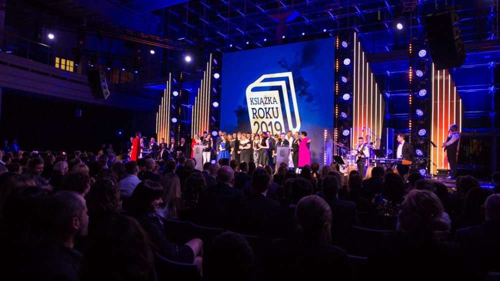 Grupa ludzi na scenie, pod sceną widownia. Za plecami laureatów duży ekran i pionowe dekoracje świetlne.