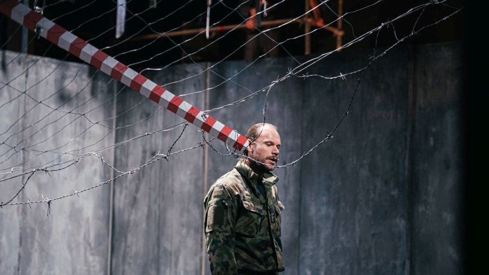 Mężczyzna w wojskowym mundurze stoi na środku sceny. Za nim betonowa ściana, nad nim zasieki i słup graniczny.