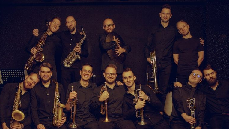 Grupowe zdjęcie dwunastu muzyków orkiestry. Mężczyźni są ubrani w ciemne koszule, każdy z nich trzyma w ręce instrument.