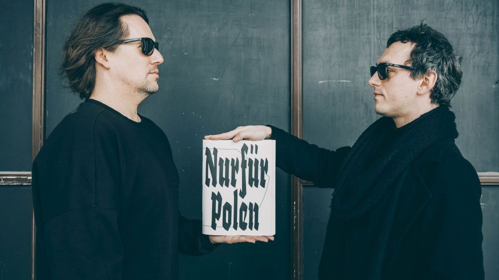Dwóch mężczyzn w czarnych ubraniach i okularach przeciwsłonecznych, zwróconych ku sobie. Wspólnie trzymają książkę z biała okładką i czarnym tytułem.
