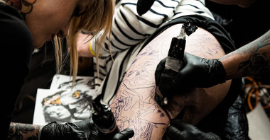 Dwoje tatuażystów tatuuje nogę leżącego chłopaka.
