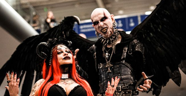 Para w gotyckich strojach stoi prezentując swoje outfity. Kobieta ma długie, czerwone włosy i czarne rogi na głowie, mężczyzna dużo kolczyków i makijaż przypominający postać filmowego Nosferatu.