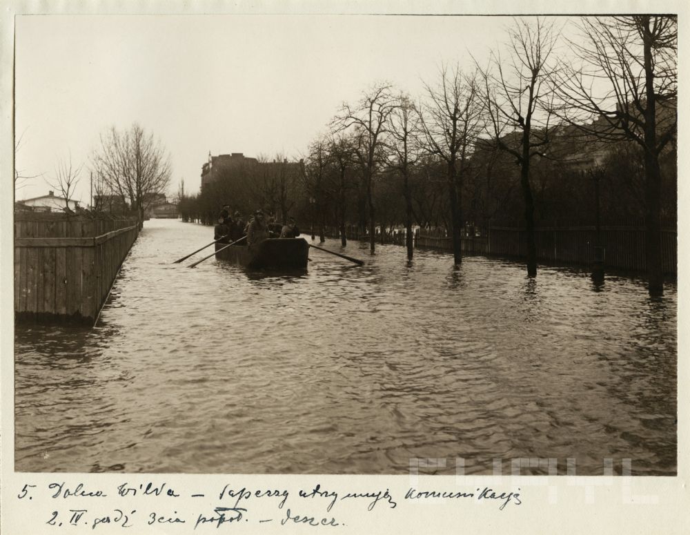 Powódź w mieście, ludzie płyną łodzią środkiem ulicy. Po prawej drzewa, po lewej płot. - grafika artykułu