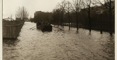 Powódź w mieście, ludzie płyną łodzią środkiem ulicy. Po prawej drzewa, po lewej płot.