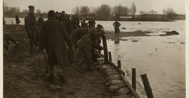 Grupa mężczyzn umacnia wał przeciwpowodizowy deskami i workami z piaskiem.