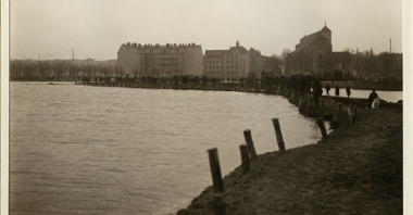 Widok na brzeg Warty, nad wodą ludzie, w tle zabudowania miejskie.