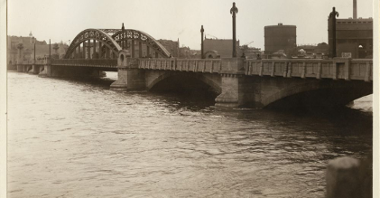 Zdjęcie mostu z dwoma półokrągłymi przęsłami. W tle budynki.