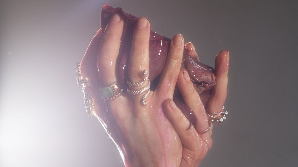 Mokre kobiece ręce w pierścionkach trzymają fragment ciała, prawdopodobnie ludzkie serce.