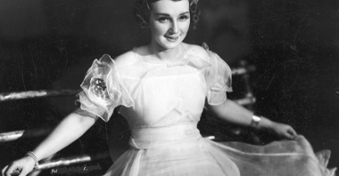 Młoda kobieta w białej sukni i z ozdobną opaską na głowie rozkłada na boki poły sukni, uśmiecha się.