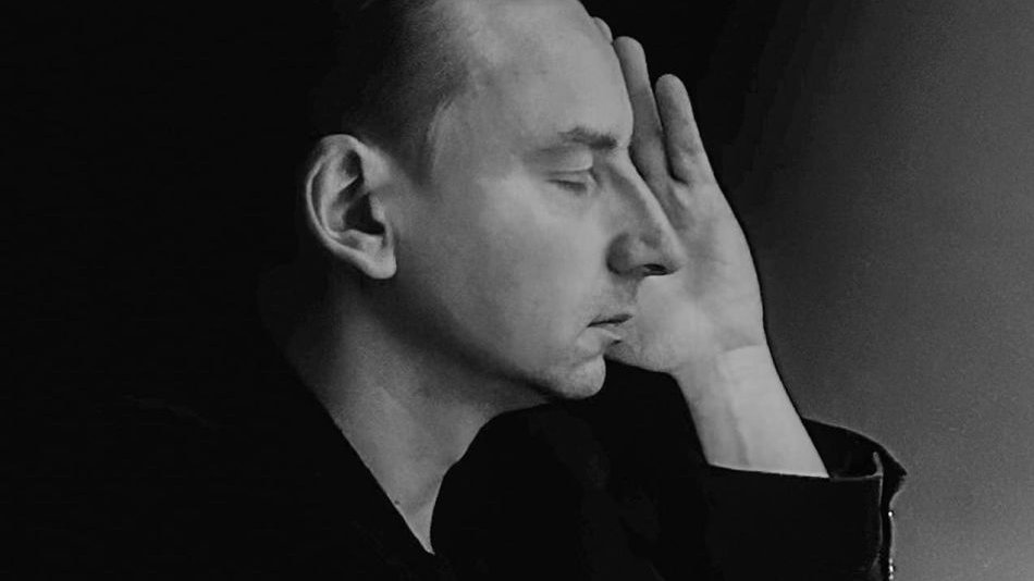 Czarno-białe zdjęcie, artysta pozuje z profilu, ma zamknięte oczy, przykłada dłoń do policzka. Wrażenie melancholijne.