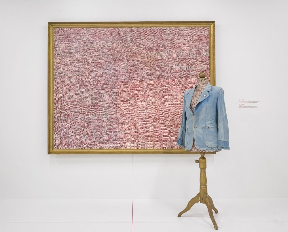 Duży obraz w ramie przypominający fakturą różowy materiał wisi na ścianie galerii. Przed obrazem stoi manekin krawiecki ubrany w dżinsową marynarkę. - grafika artykułu