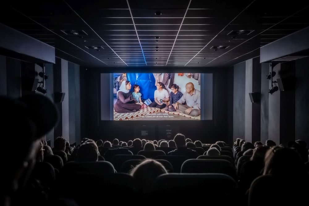 Widzowie w kinie oglądają film na dużym ekranie. Jest ciemno, salę rozświetla tylko światło z ekranu. - grafika artykułu