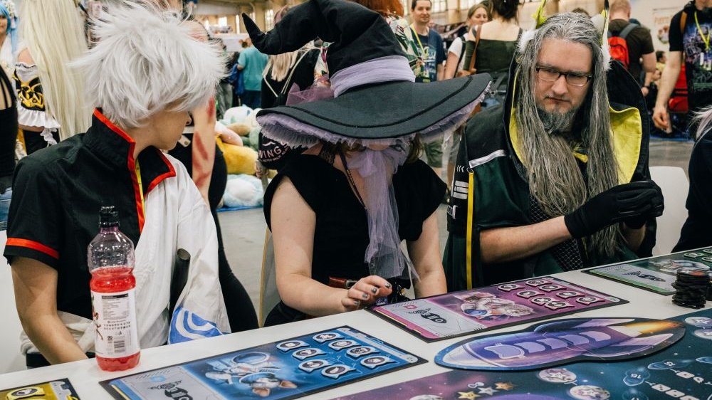 Trzy osoby przebrane w postaci ze świata anime i fantasy siedzą przy stole i grają w planszówkę. W środku siedzi postać w czarnej sukience i wielkim czarnym kapeluszu wiedźmy na głowie.