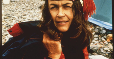 Wanda Rutkiewicz w górskim stroju i chuście z frędzlami na głowie pozuje na tle rozbitego namiotu pod zdobywanym szczytem.