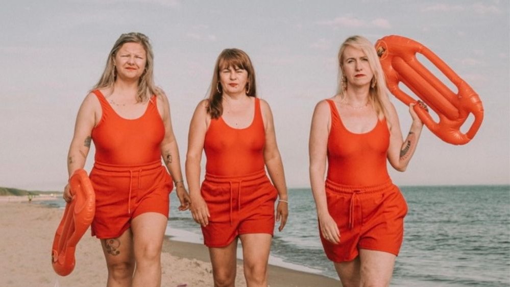 Trzy kobiety idą plażą w jaskrawo pomarańczowych strojach ratowniczek wodnych.
