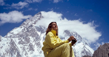 Wanda Rutkiewicz w żółtym kombinezonie i z aparatem w rękach siedzi na kamieniu na tle ośnieżonej góry.