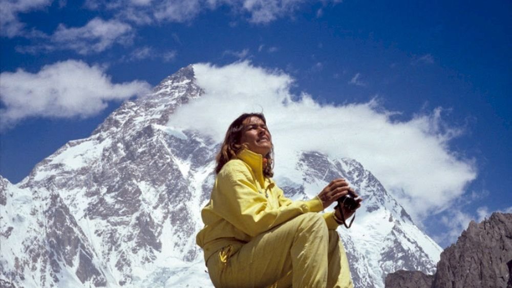 Wanda Rutkiewicz w żółtym kombinezonie i z aparatem w rękach siedzi na kamieniu na tle ośnieżonej góry.