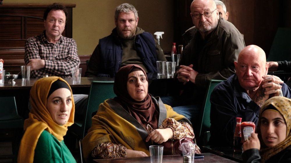 Grupa kobiet w muzułmańskich chustach i mężczyzn siedzi przy stołach. Wszyscy mają poważne miny.