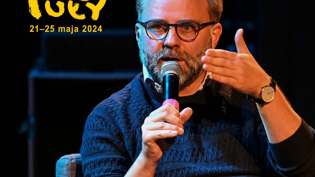 Mężczyzna w okularach siedzi na krześle, mówi do mikrofonu, gestykuluje ręką. W rogu zdjęcia logo Festiwalu Fabuły