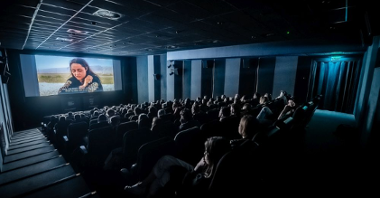Ekran kinowy w zaciemnionym kinie, widać głowy publiczności oglądającej film