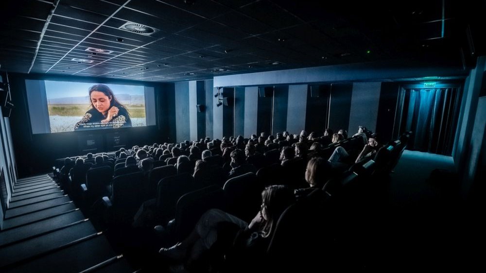 Ekran kinowy w zaciemnionym kinie, widać głowy publiczności oglądającej film